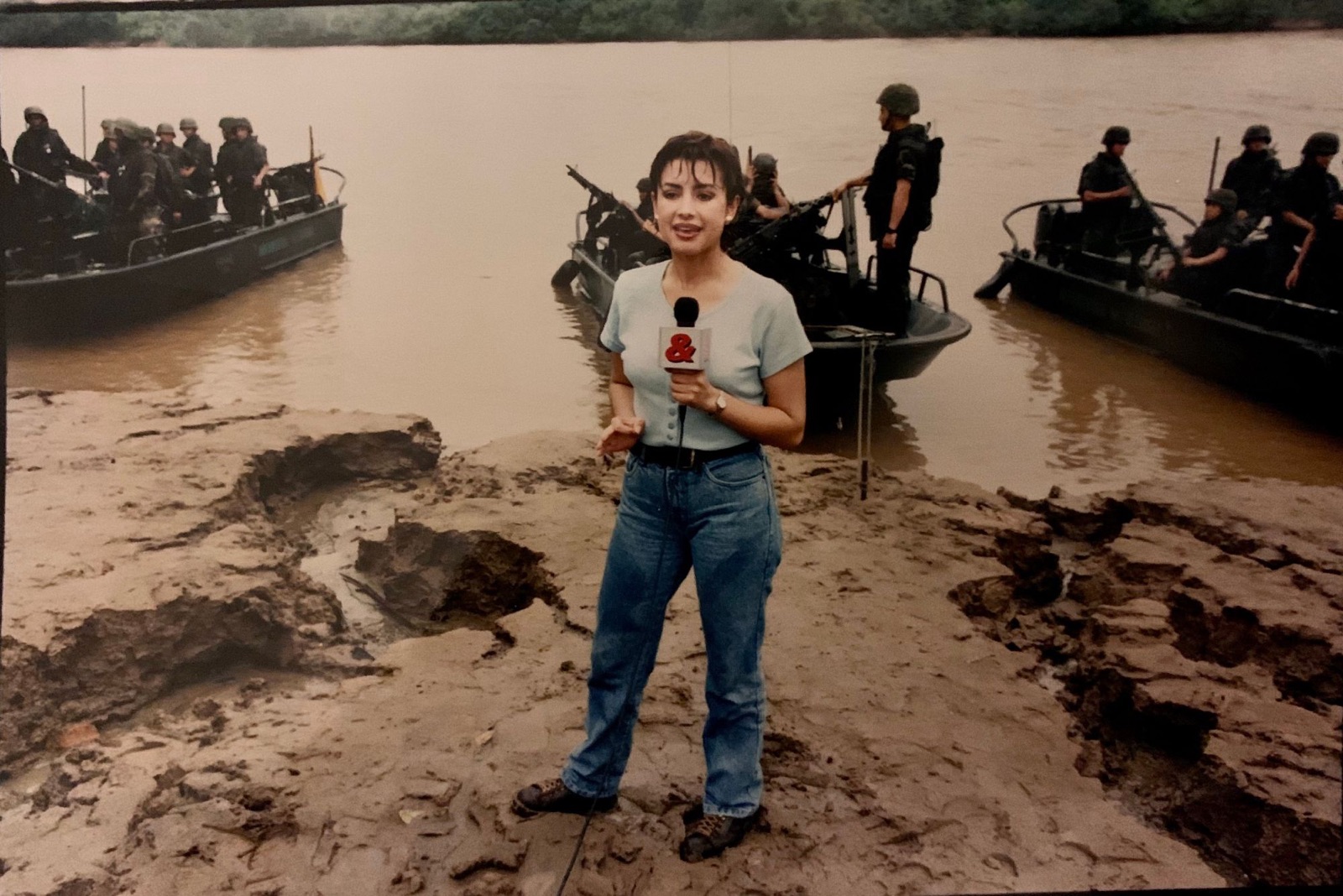 Adriana Aristizábal reportea para RCN Televisión a orillas de un río en Colombia, con lanchas militares armadas detrás